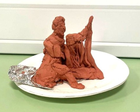 A small clay sculpture of a person in a long cloak playing a harp. It sits on a paper plate.