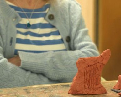 A sculpture of a harp in terracotta clay. Behind it we can see a person with their arms crossed, sitting at the table.