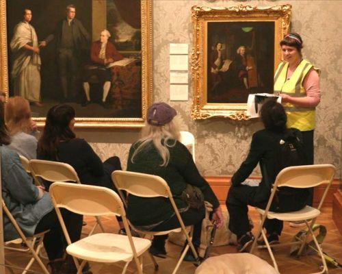A group of people sitting on plastic chairs in an art gallery. Someone is standing in front of them, in a hi-vis jacket, talking about the art on the walls.