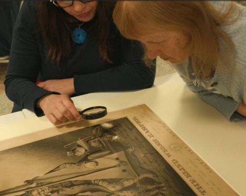 Two women leaning against a table, looking closely at a black and white print of a harpist. One of them is holding a magnifying glass to the image.