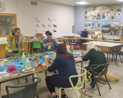 Four women gathered around a table in a colourful art activity room, smiling and making clay sculptures.